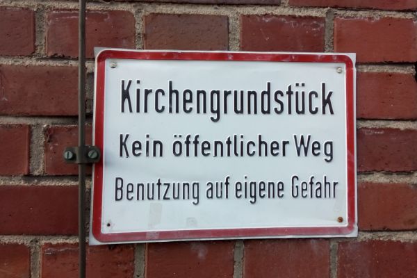 Ein Schild an einer Wand mit der Aufschrift: Kirchengrundstück. Kein öffentlicher Weg. Benutzung auf eigene Gefahr.